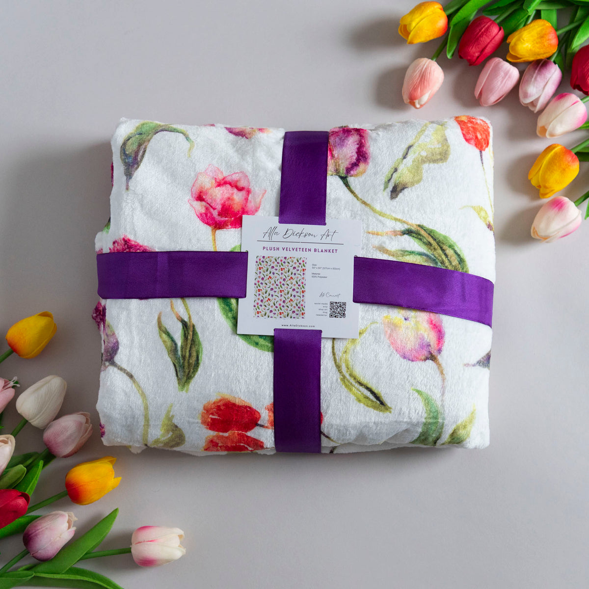 A folded white plush blanket with a colorful watercolor tulip print is wrapped in a purple ribbon and a white paper tag, styled on a light gray surface with decorative tulips in the corners.