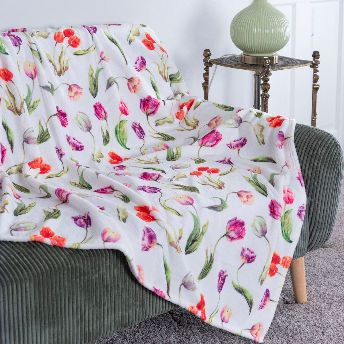 A white throw blanket with a colorful watercolor tulip print is draped over a green corduroy armchair in a bright room, with a small table and green vase in the background.
