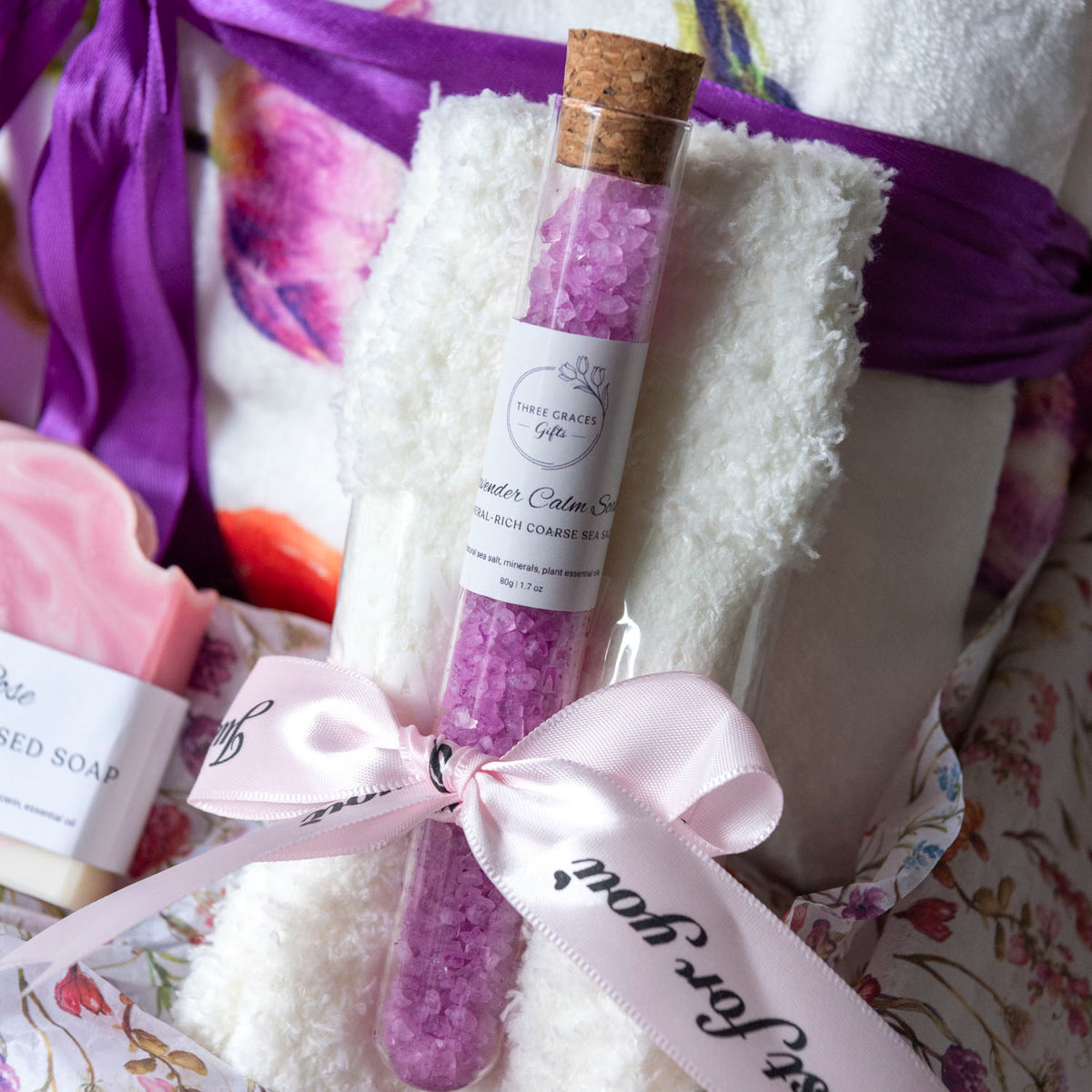 A gift set featuring a clear glass tube of purple bath salts with a cork stopper, wrapped in a light pink ribbon. The tube is placed on a white fluffy towel, surrounded by other gift items including a pink soap bar and purple ribbons, all set against a floral fabric background.