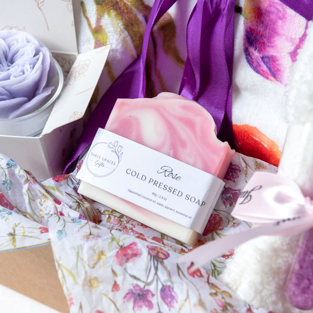 A pink and white swirled bar of Rose Cold Pressed Soap by Pure Grace Soap nestled in floral tissue paper inside a gift box, alongside a rose candle
