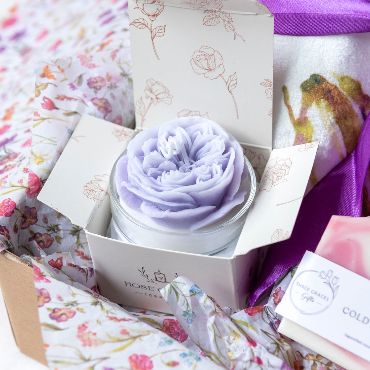 A lavender-colored sculpted soap or candle in a clear glass jar, presented inside an open white box with floral linework and purple accents, surrounded by matching floral tissue paper and gift packaging.
