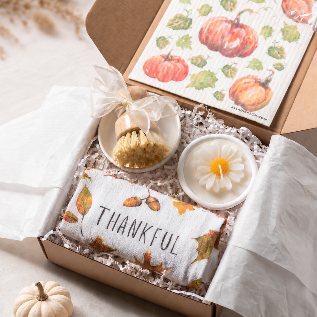An open kraft gift box filled with fall-themed items, including a printed thankful tea towel, a daisy flower candle, a bamboo scrubber and ceramic holder dish, and a pumpkin swedish dishcloth styled on a light surface with small decorative pumpkins.