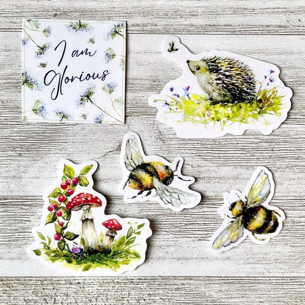 A set of five die-cut stickers featuring nature illustrations, including two bumblebees, a hedgehog in a flower patch, a mushroom cluster with red berries, and a square card reading 'I am Glorious', arranged on a light gray wood plank background.