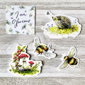 A set of five die-cut stickers featuring nature illustrations, including two bumblebees, a hedgehog in a flower patch, a mushroom cluster with red berries, and a square card reading 'I am Glorious', arranged on a light gray wood plank background.