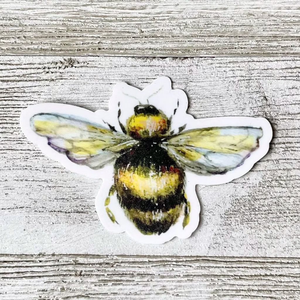 A die-cut sticker of a yellow and black bumblebee with translucent wings, placed on a light gray wood plank background.