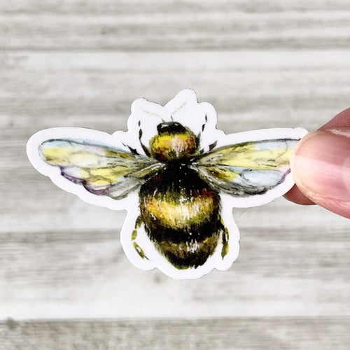 A hand holding a die-cut sticker of a yellow and black bumblebee against a light gray wood plank background.