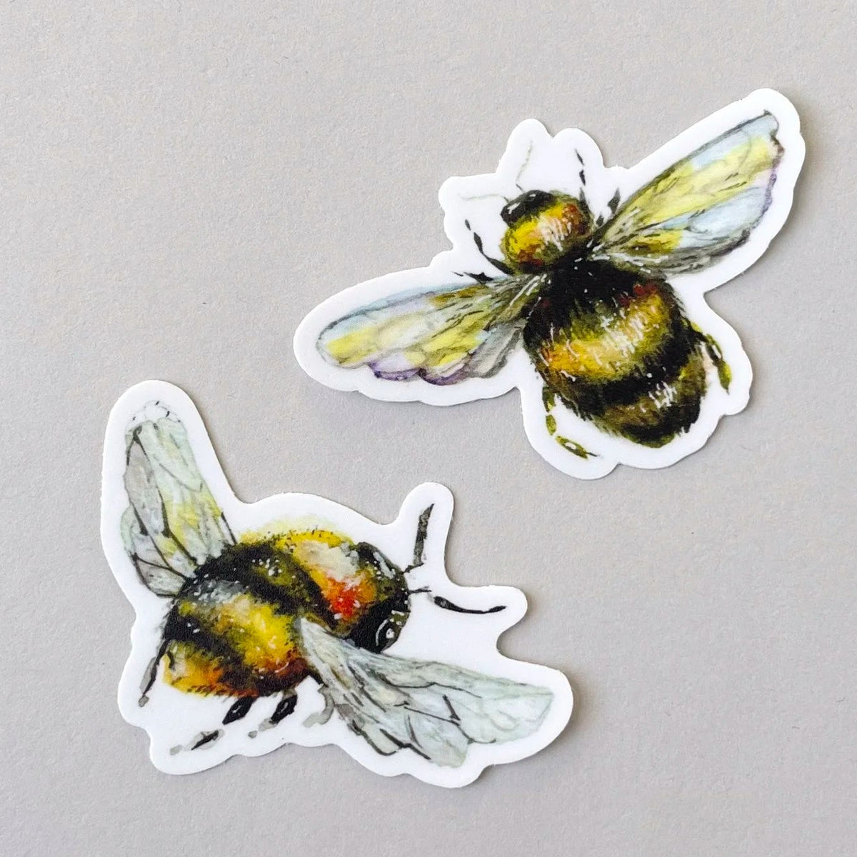 Two die-cut stickers featuring watercolor-style bumblebees in yellow, black, and white, displayed on a neutral light gray background.