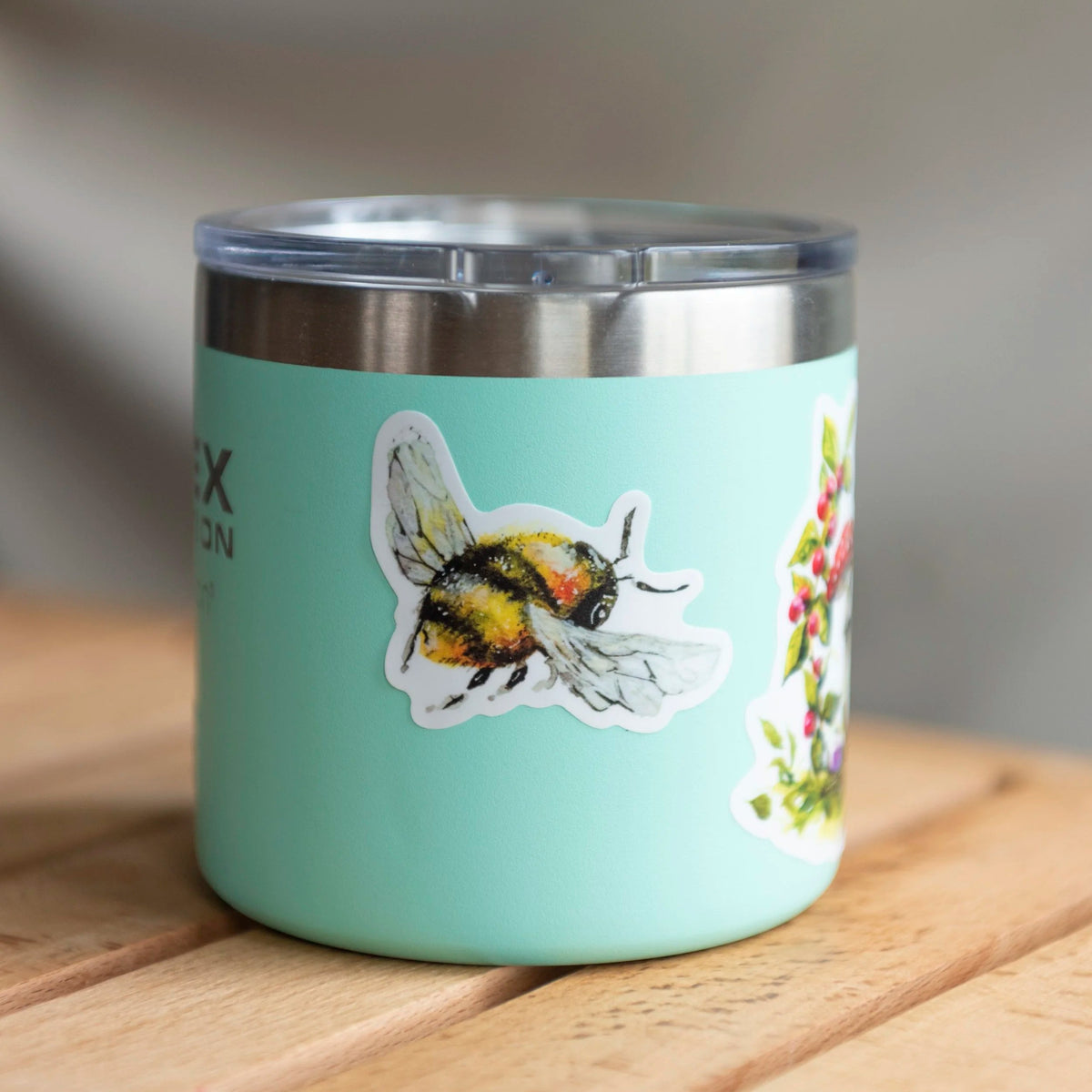 A mint green insulated tumbler with a clear lid sits on a wooden surface. The tumbler is decorated with a die-cut sticker of a bumblebee and a partial floral sticker on the right side.