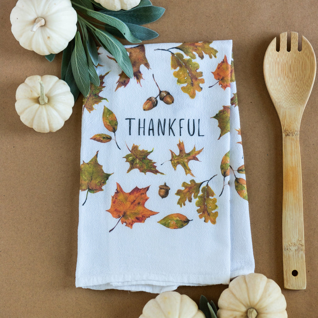 A white kitchen tea towel with words 'THANKFUL' and autumn leaves printed on it, styled in a flat lay on a brown background with small white pumpkins, greenery, and a wooden spoon.